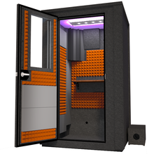 Load image into Gallery viewer, Angled view of the WhisperRoom&#39;s Voice Over Deluxe Package - a 4&#39; x 4&#39; double-wall vocal booth, thoughtfully equipped with acoustic treatment, a functional desk, studio lighting, and an array of premium features. The left-hinged door is open, revealing the interior enhanced by vibrant orange StudioFoam, creating an ideal recording atmosphere.
